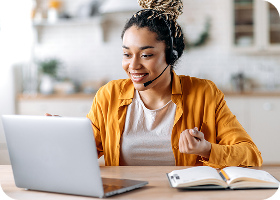 Jeune femme afro-américaine sympathique et attirante portant un casque, employée d'un centre d'appels, opératrice de support, travaillant sur un ordinateur portable, effectue une consultation en ligne avec un client, regardant l'écran, souriant