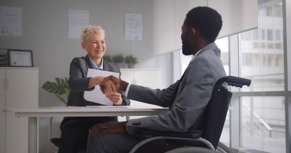 Un candidat africain handicapé et une employeuse mature se serrent la main et sourient à la caméra. Un candidat afro-américain en fauteuil roulant serre la main de son responsable RH.